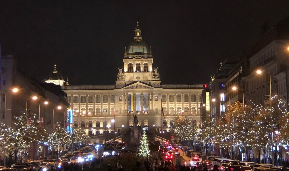 A grand historic building illuminated at night, with a large Christmas tree and festive street lights lining the busy avenue leading up to it.