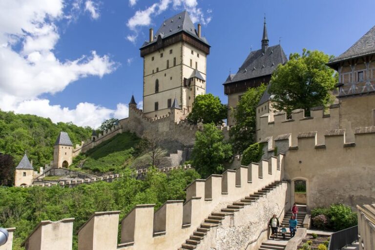 A steep stone staircase leads up to Karlštejn Castle, a Gothic fortress surrounded by lush greenery and set against a bright blue sky with scattered clouds. The castle's tall central tower and fortified walls dominate the landscape, showcasing medieval architecture in the Czech countryside.