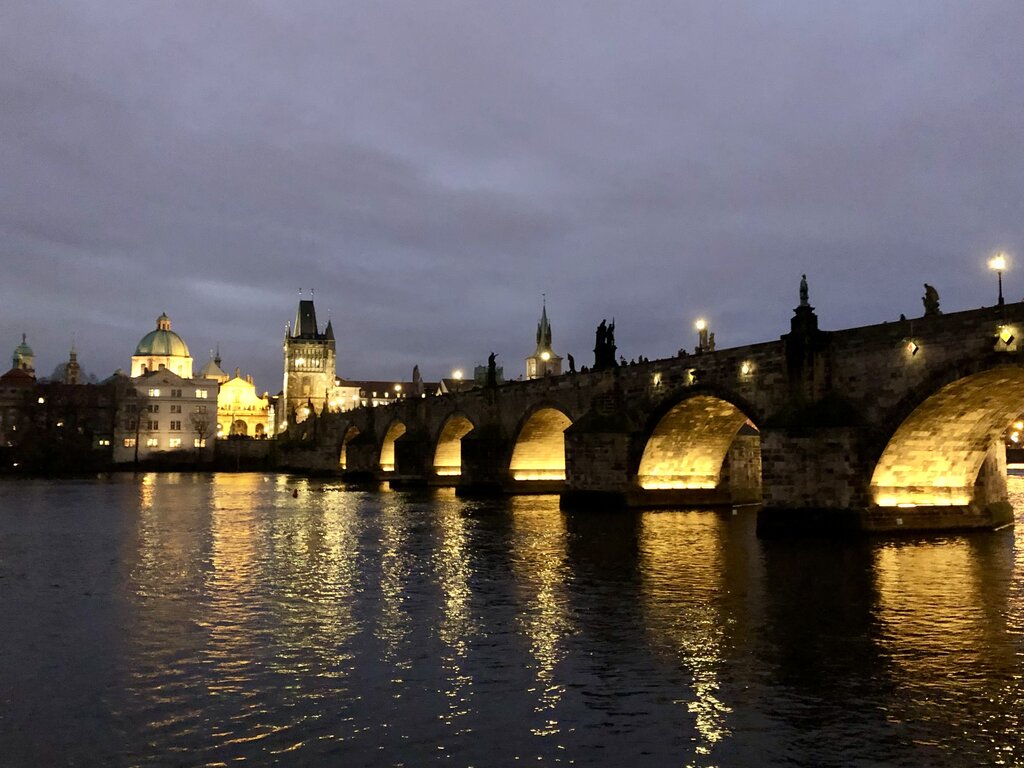 The Charles Bridge in Prague glows with golden lights reflecting on the Vltava River at dusk, with the Old Town skyline softly illuminated in the background.