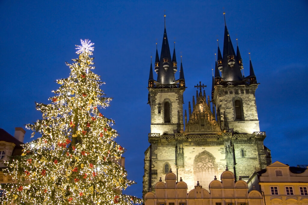 The Church of Our Lady before Týn towers over Old Town Square beside a sparkling Christmas tree covered in golden lights against a deep blue evening sky.