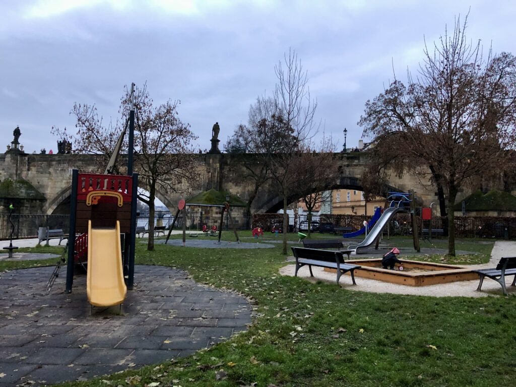 Children’s playground near Charles Bridge in Prague with empty swings, a yellow slide, and a sandbox where a toddler in a striped hat is playing. Bare trees and cloudy skies suggest a chilly autumn or winter day.