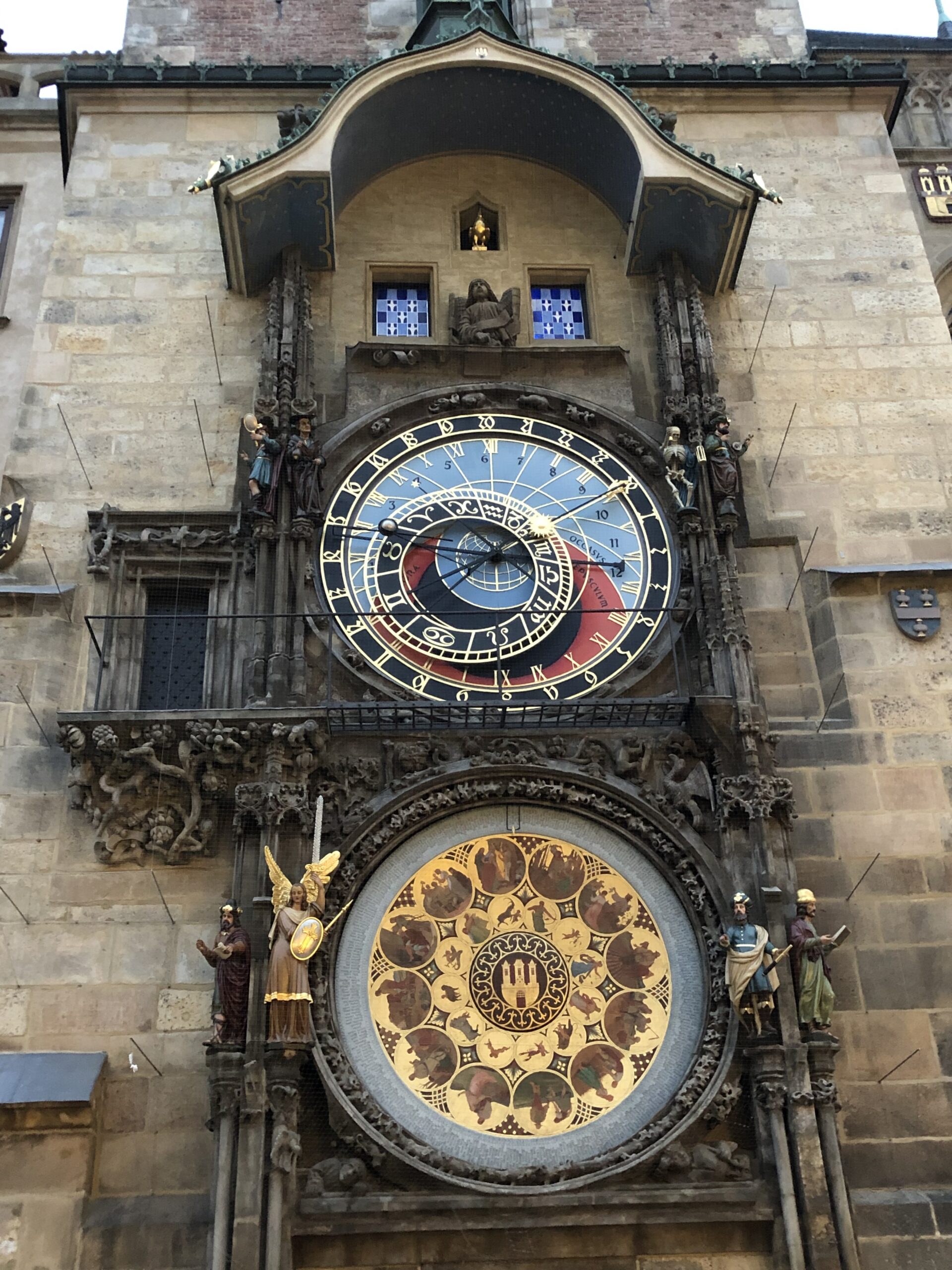 Close-up view of the Prague Astronomical Clock, or Orloj, showing its ornate details including zodiac signs, golden Roman numerals, and mechanical figurines. The centuries-old clock is set into a stone wall and decorated with intricate carvings.