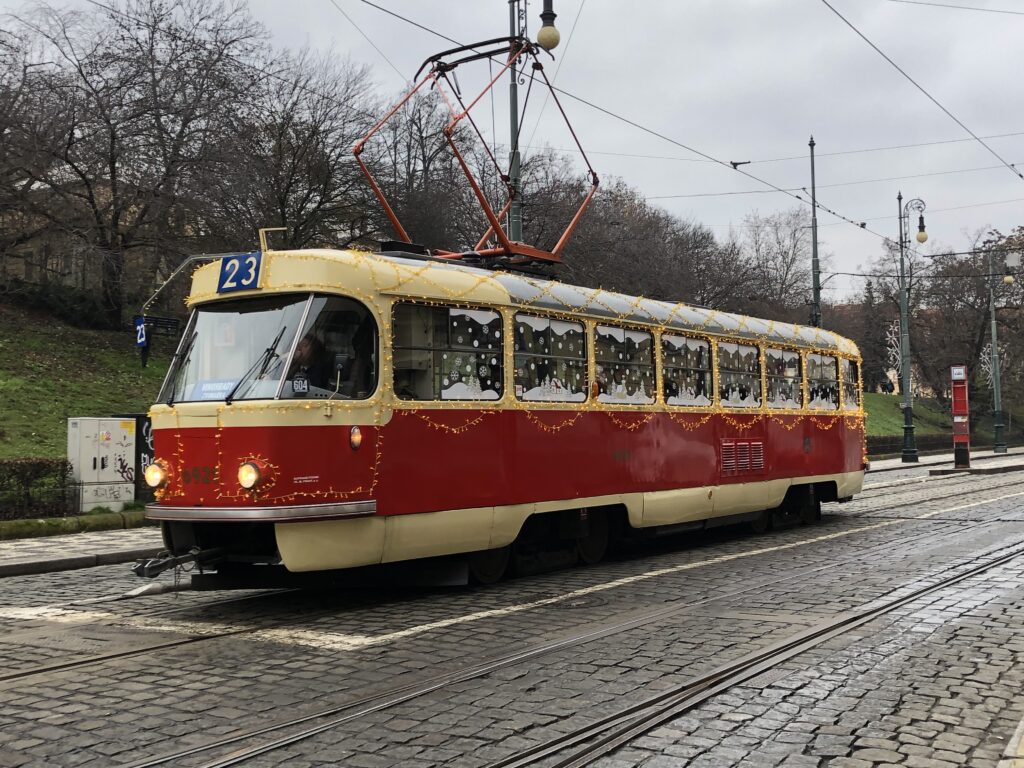 A vintage red and cream tram on route 23 in Prague is decorated with festive Christmas lights and snowflake decals on the windows. The tram travels along cobblestone tracks on an overcast winter day, with bare trees and classic street lamps lining the route.