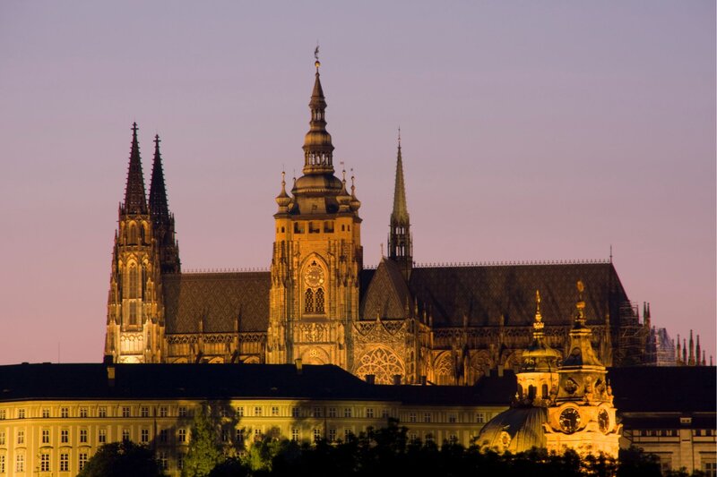 St. Vitus Cathedral in Prague glows with warm lighting at dusk, its Gothic spires and ornate facade standing tall above the surrounding buildings. The sky fades into soft shades of pink and purple, highlighting the cathedral’s dramatic architecture.
