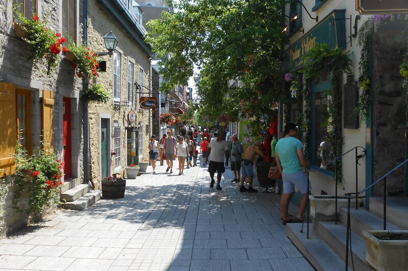 A lively cobblestone street in Old Quebec lined with stone buildings, flower boxes, and boutique shops, with tourists strolling and browsing on a sunny day. The street feels quaint and European with its historic charm and colorful details.