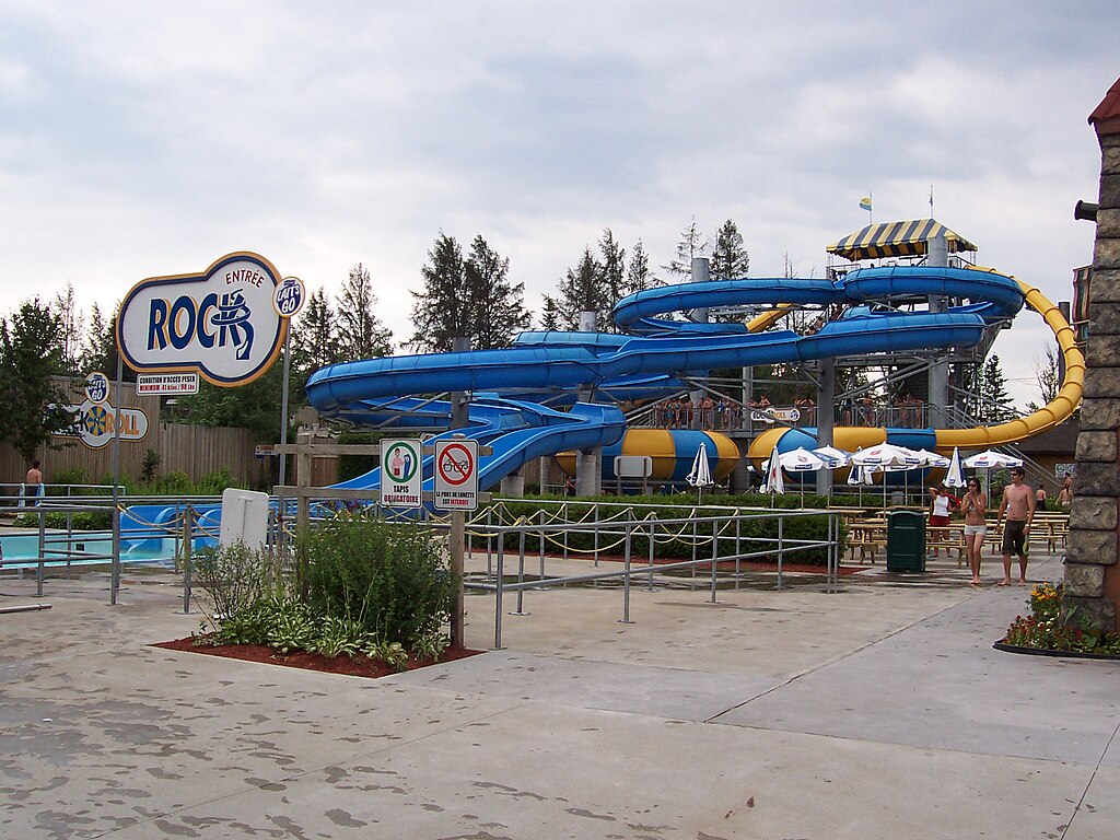 A colorful water park slide complex featuring twisting blue and yellow tube slides supported by metal scaffolding. The foreground shows a sign for the ride "Roc-N-Roll" and a queue area with wet pavement and a few guests in swimsuits walking nearby under a cloudy sky.
