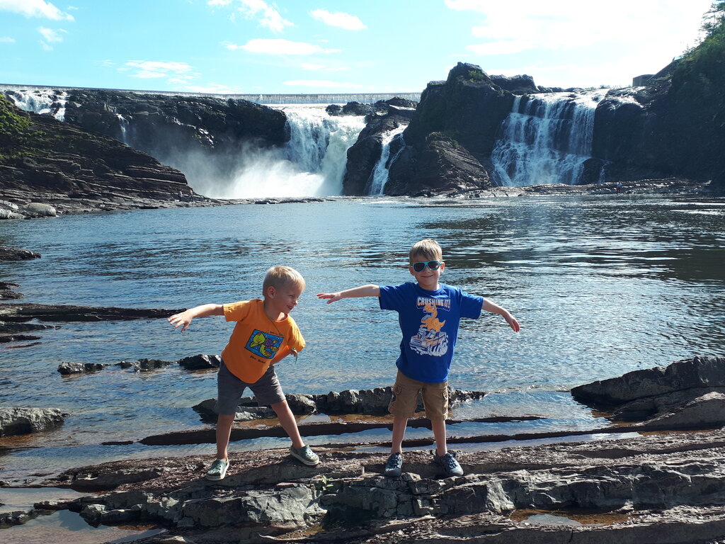 Two young boys pose playfully on rocky terrain in front of the Chutes de la Chaudière waterfalls near Quebec City. The powerful falls cascade over dark cliffs into a wide, calm river, with a pedestrian suspension bridge visible in the background under a bright blue sky.