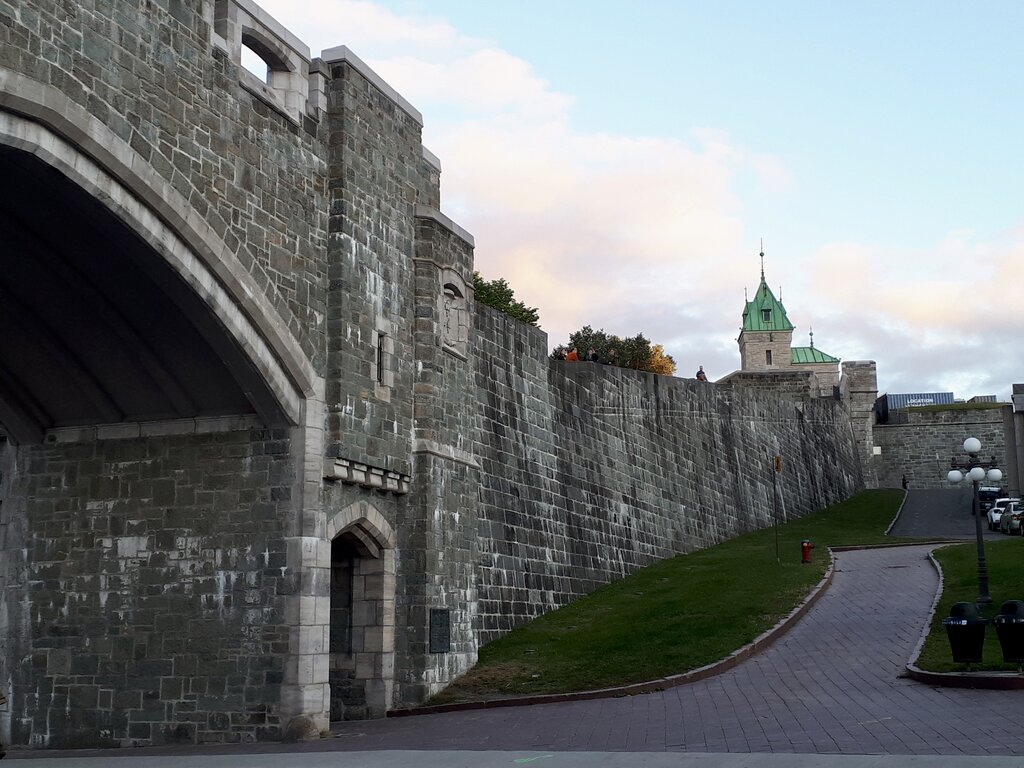A view of the fortified stone walls of Old Quebec at dusk, showcasing one of the historic city gates with a green copper-roofed tower rising in the distance. A winding path and road lead up beside the wall, where a few visitors are walking along the top. The sky is soft with pastel clouds, highlighting the city's rich architectural heritage.