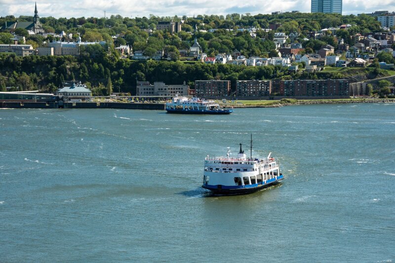 A ferry crosses the St. Lawrence River between Quebec City and Lévis, with the cityscape and green hills of Lévis visible in the background. The water shimmers under a partly cloudy sky, capturing a scenic and peaceful view of the Quebec-Lévis ferry service.