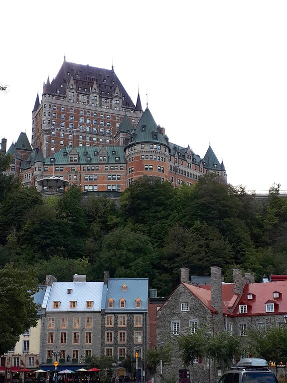 A large castle-like hotel with green rooftops rises above colourful historic stone houses nestled at the base of a hill covered in trees. This is the Chateau Frontenac in Quebec City