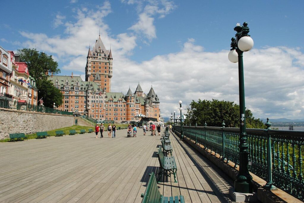 People stroll along a wide wooden boardwalk lined with green benches and vintage street lamps, leading toward a grand historic hotel overlooking the river in Quebec City