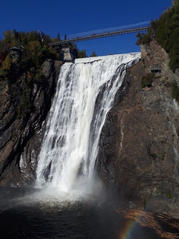 Montmorency Falls near Quebec City, with water cascading down a cliff and a pedestrian suspension bridge above, framed by autumn trees and a visible rainbow.