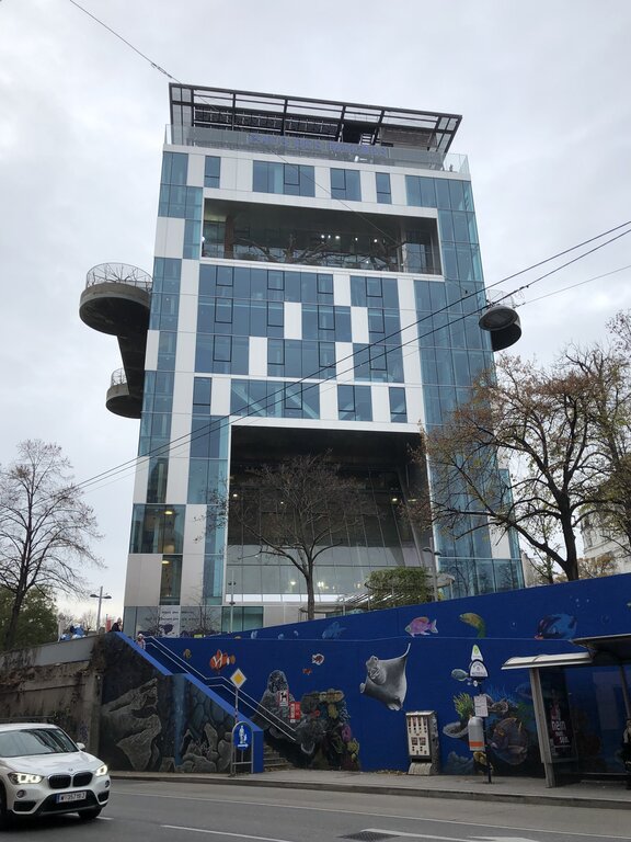 A modern glass building known as Haus des Meeres in Vienna towers above a street, with a blue mural of sea creatures wrapping around the entrance and a spiraling outdoor staircase on the left.