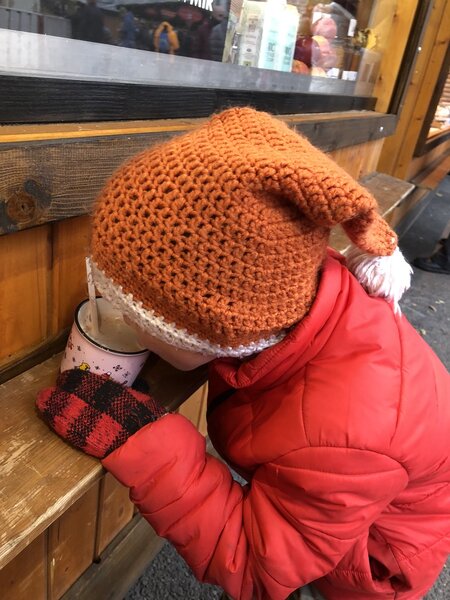 A young child in a red winter coat and orange knit hat with a white pom-pom drinks from a festive mug while leaning on a wooden bench outside a cozy market stall. The child wears red and black plaid gloves, adding to the warm holiday atmosphere.