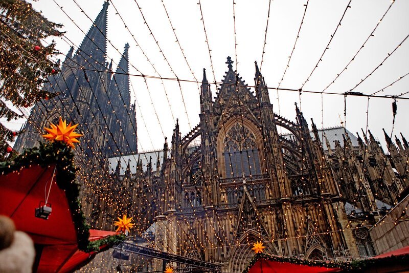 View of Cologne Cathedral's ornate Gothic facade framed by strings of festive Christmas lights and glowing star-shaped decorations above red market stalls during the holiday season.