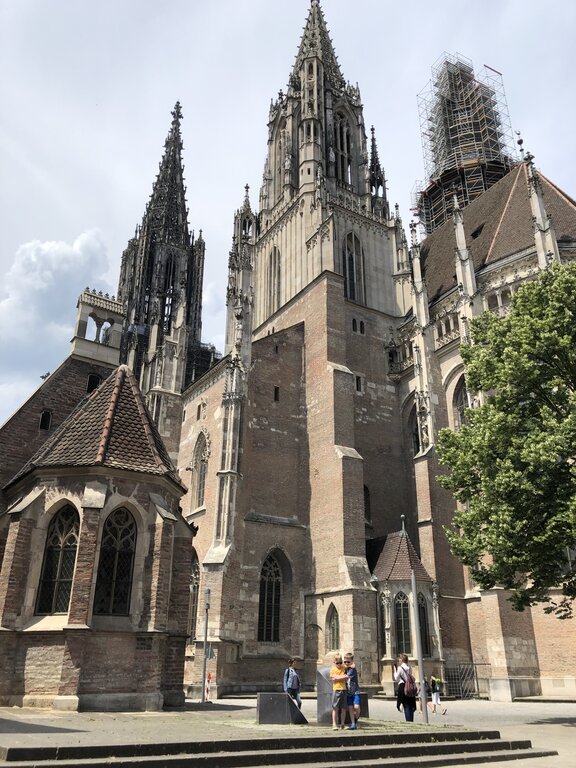 Two boys stand at the base of Ulm Minster, a towering Gothic cathedral with intricate spires and scaffolding on one tower, set against a partly cloudy summer sky.