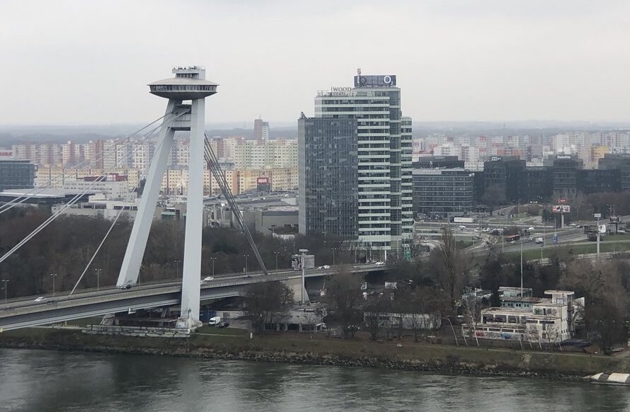 The futuristic UFO Bridge spans the Danube River, featuring a flying saucer-like observation deck atop its tower. The modern cityscape with high-rises and residential blocks stretches across the background.