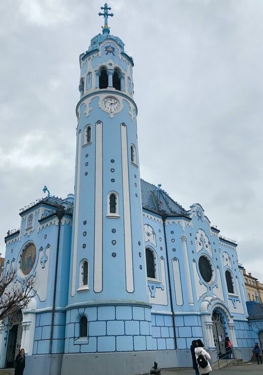 A pastel blue Art Nouveau church with intricate white detailing and a tall clock tower topped with a blue cross, standing out under an overcast sky. Visitors walk past its whimsical facade and curved archways in Bratislava, Slovakia