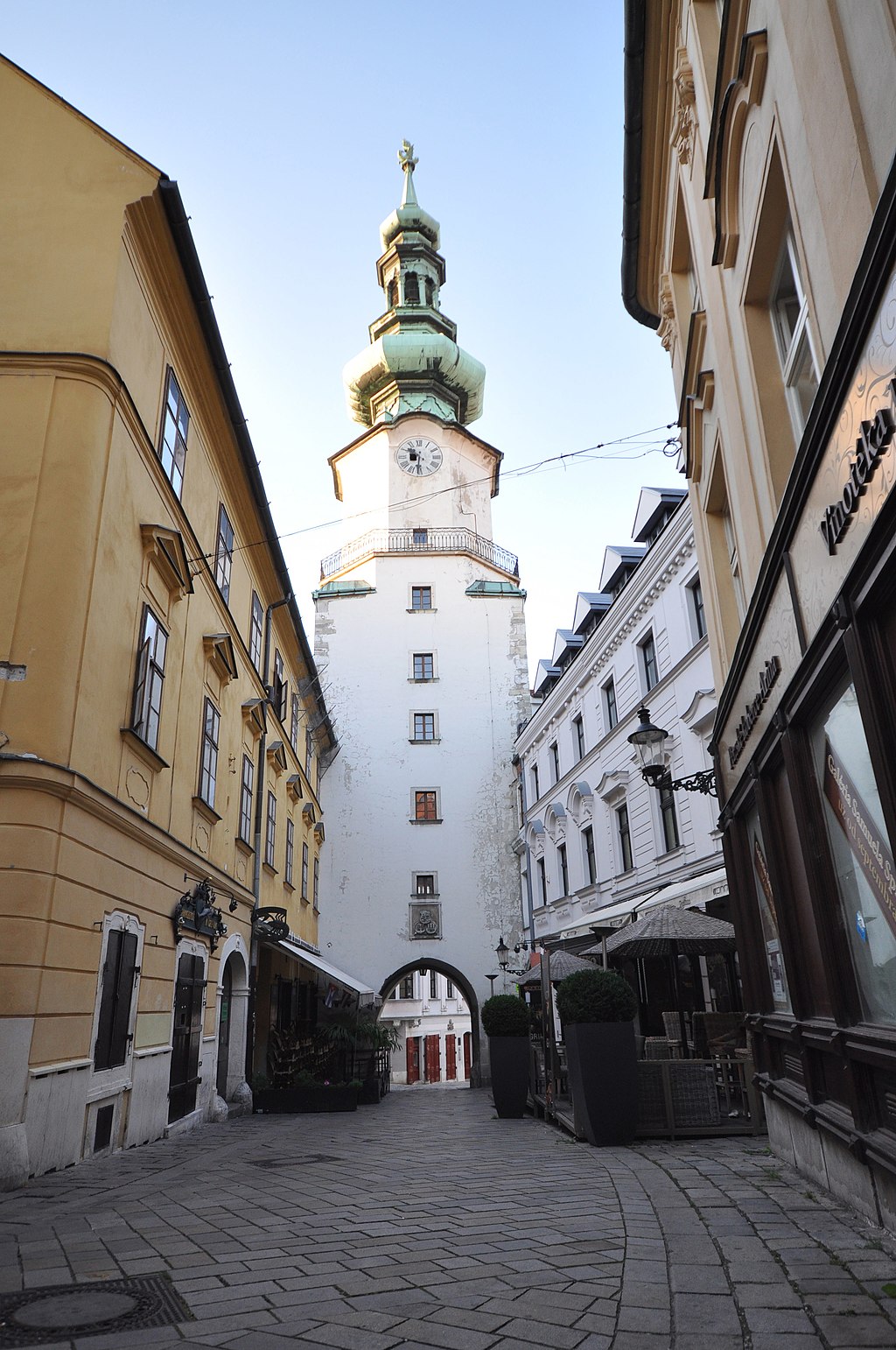 Michael’s Gate, a tall white medieval tower with a green copper spire and clock, rises between narrow cobblestone streets lined with historic buildings in Bratislava’s Old Town.