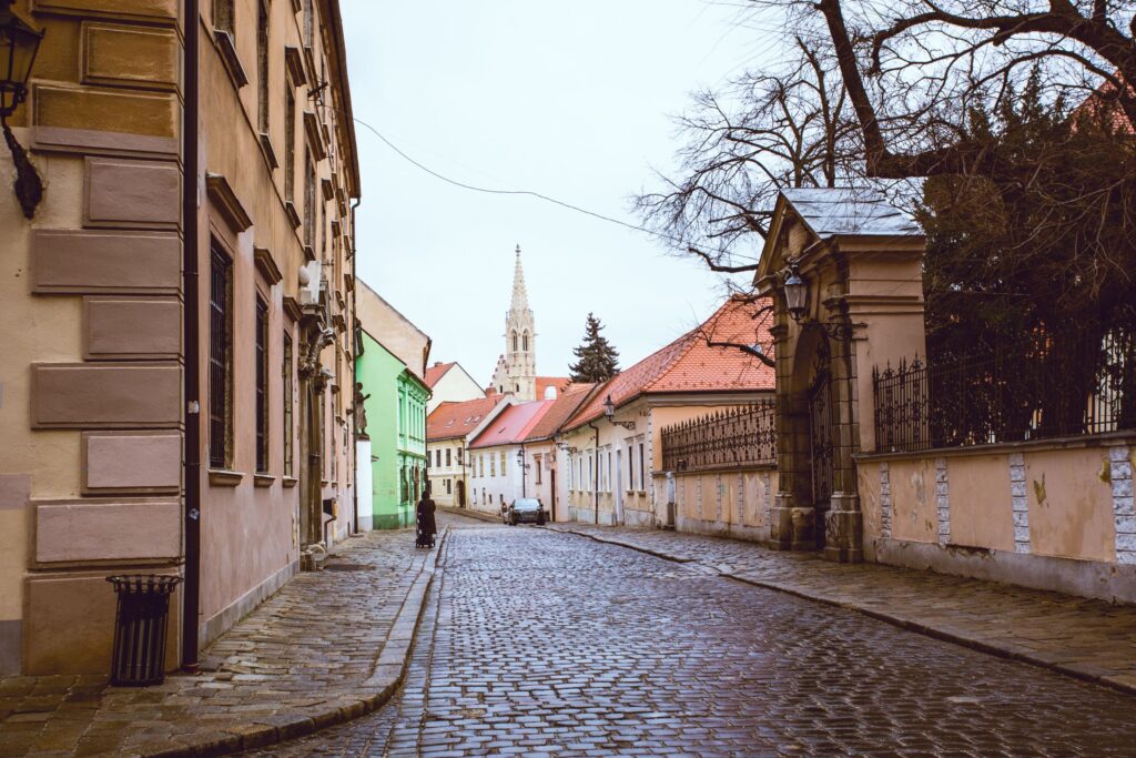 A quiet, winding cobblestone street in Bratislava’s Old Town is lined with pastel-colored buildings and leads to a distant Gothic church tower under an overcast sky.