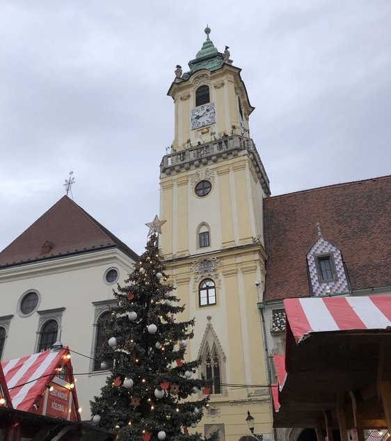 A historic yellow clock tower rises above red-and-white striped market stalls and a decorated Christmas tree in Bratislava’s main square. The overcast sky adds a wintry feel to the festive holiday setup.