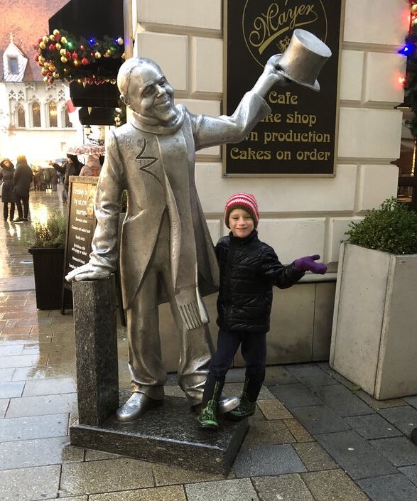 A silver statue of Schöner Náci, a well-known local figure, tips his top hat with a friendly smile as a child poses beside him on a rainy street in Bratislava.