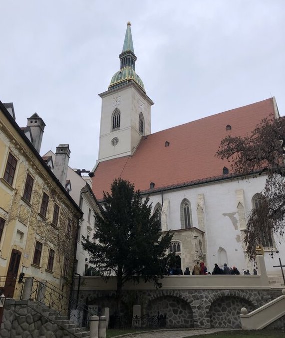 A tall white Gothic cathedral with a green spire and red-tiled roof stands against a cloudy sky, surrounded by narrow stone streets and old buildings. A group of visitors is gathered at the cathedral’s entrance.