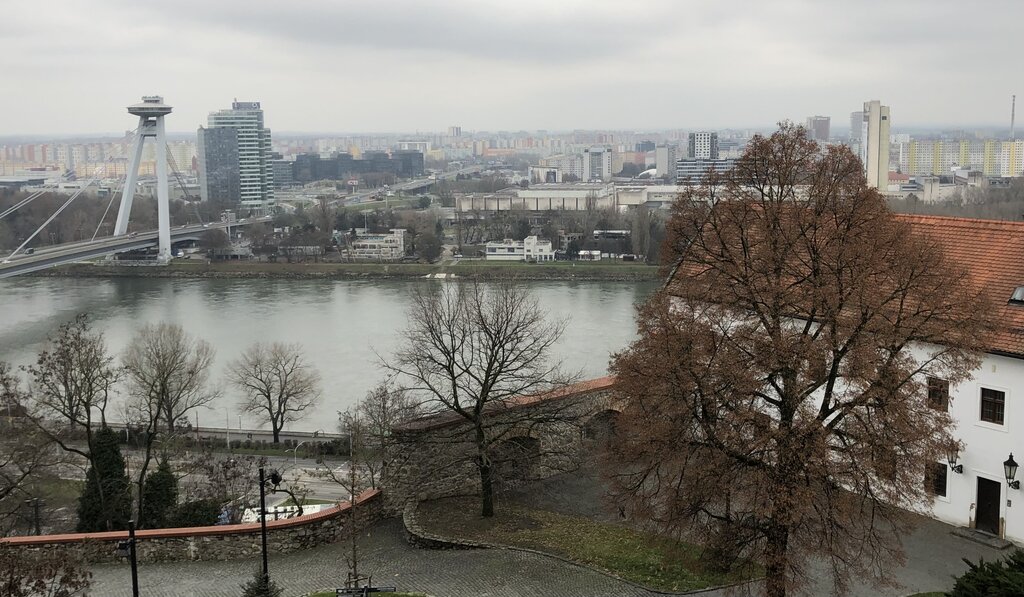 A panoramic view from Bratislava Castle shows the UFO Bridge spanning the Danube River, with city buildings and colourful apartment blocks stretching into the distance under gray skies.
