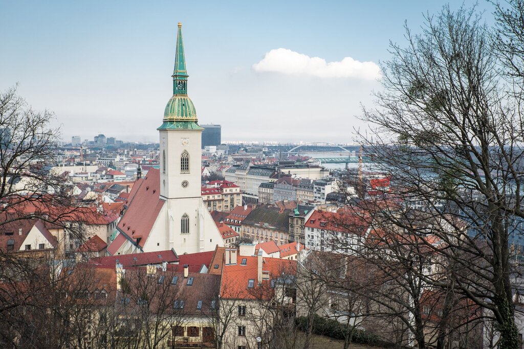 A scenic cityscape of Bratislava shows St. Martin’s Cathedral with its green spire rising above a sea of red rooftops and modern buildings, with the Danube and bridges in the distance.