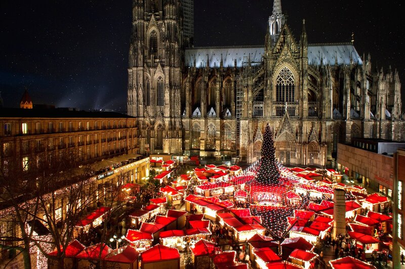 A festive Christmas market at night, set beneath the towering Cologne Cathedral. Red-roofed stalls fill the square, centred around a tall Christmas tree wrapped in strings of white lights that spread outward like a canopy. Crowds of people walk through the glowing market surrounded by Gothic architecture.