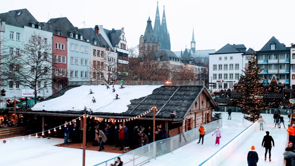 People ice skate at an outdoor rink surrounded by historic buildings and a decorated Christmas tree, with the Cologne Cathedral visible in the background.
