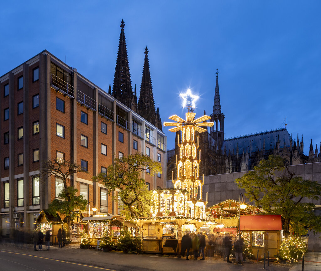 Brightly lit Christmas pyramid and cozy wooden stalls glow under the evening sky at Adventsdorf am Hof, with the spires of Cologne Cathedral rising in the background.