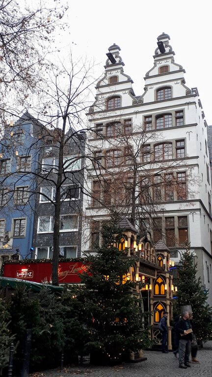 Tall historic buildings line a street filled with decorated Christmas trees and a glowing wooden market stall, creating a festive winter scene.