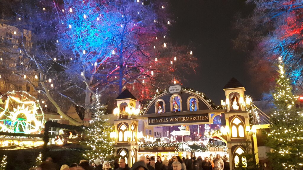 The illuminated entrance arch to "Heinzels Wintermärchen" Christmas market in Cologne, with festive buildings, decorated trees, and colourful lights in the background.