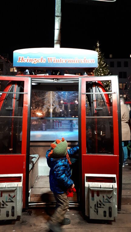 A child in winter clothes steps into a red gondola cabin at the Heinzels Wintermärchen market in Cologne Germany, with festive lights and people in the background.