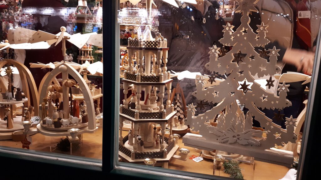 A market stall window filled with handcrafted wooden Christmas decorations, including candle arches and a tall Christmas pyramid, with shoppers browsing in the background.