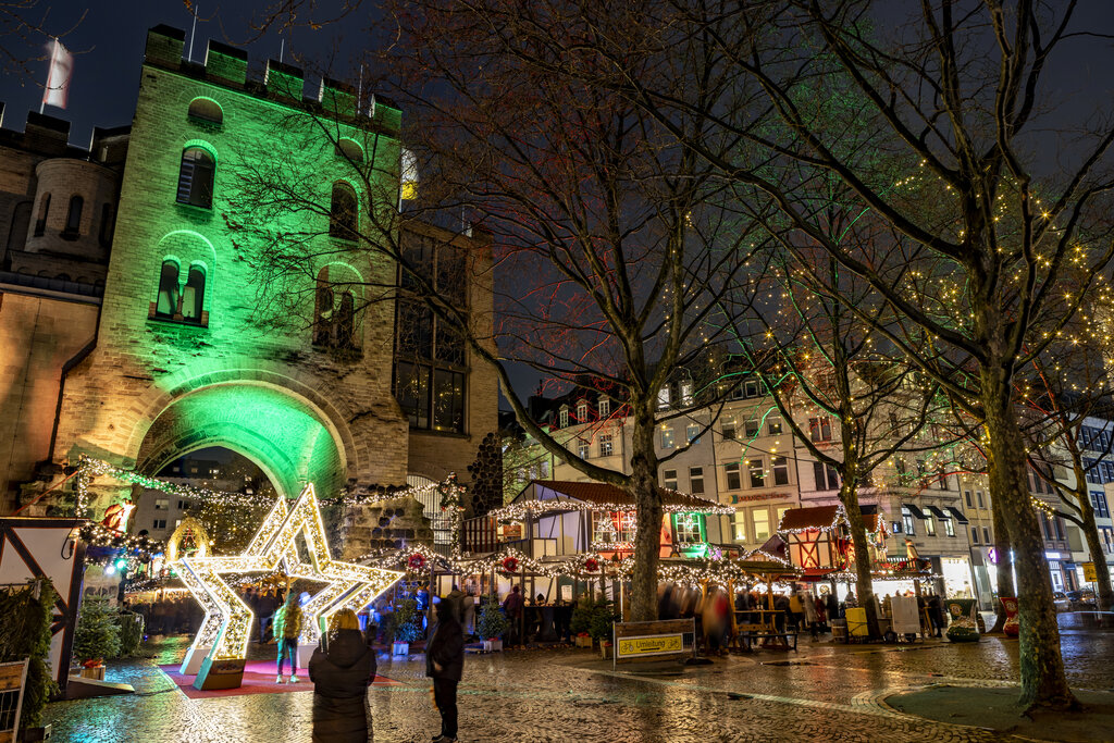 Festive night scene of Rudolfplatz in Cologne, with a medieval-style gate illuminated in green and a glowing star installation marking the entrance to a charming Christmas market lined with twinkling trees and decorated stalls.