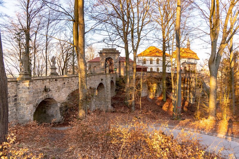 A historic stone bridge with arched supports and statues along its balustrade leads to a baroque-style chateau with a red-tiled roof, nestled among tall, leafless trees in Bohemian Paradise, Czechia. Warm golden sunlight filters through the forest, casting long shadows on the forest floor covered in dry autumn leaves.