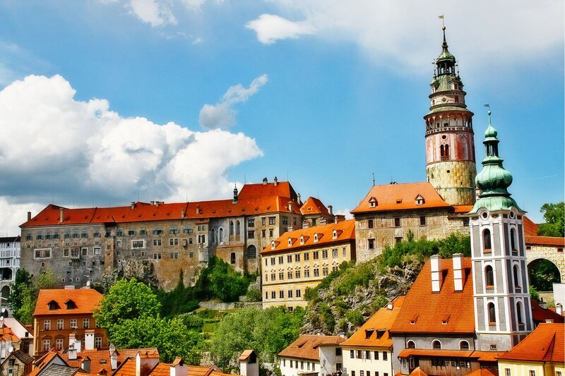 Český Krumlov Castle rises above a cluster of red-roofed buildings, with its tall, decorative tower featuring green accents and Renaissance frescoes. The historic complex sits atop a rocky hill, surrounded by greenery and set against a bright blue sky with large, billowing clouds.