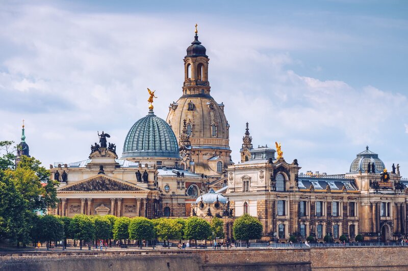 An ornate cluster of baroque and neoclassical buildings in Dresden Germany features grand domes topped with golden statues, intricate facades, and sculptural details. In the foreground, rows of trimmed trees line a riverside promenade where people stroll beneath a soft, partly cloudy sky.