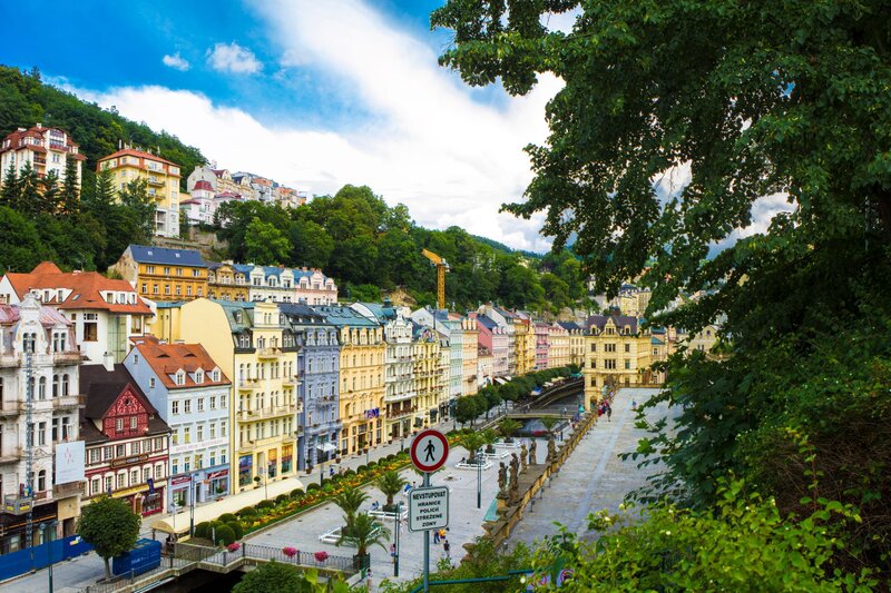 A row of colourful, ornate buildings lines a pedestrian promenade in the spa town of Karlovy Vary, with forested hills rising steeply behind them. Elegant colonnades, palm trees, and a wide walkway stretch alongside a river, creating a vibrant yet tranquil urban scene beneath a partly cloudy sky.