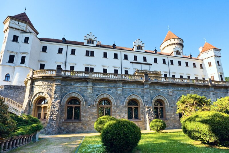 A grand historic chateau with white upper walls and a stone lower level stands against a clear blue sky, featuring arched windows, stepped gables, and conical red-roofed towers. Manicured green shrubs and a tidy garden line the foreground, adding contrast to the building’s symmetrical, fortress-like structure.