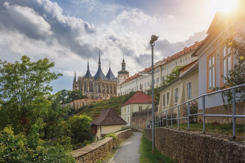 A narrow stone path winds uphill between charming houses and a historic stone wall, leading toward the towering spires of St. Barbara’s Church in Kutná Hora. The late afternoon sun breaks through dramatic clouds, casting a warm glow over the Gothic cathedral and surrounding Baroque buildings.