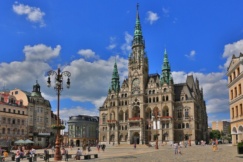 People stroll across a cobblestone square in front of the ornate Liberec Town Hall, a Neo-Renaissance building with green spires, a central clock tower, and arched windows. The sky is bright blue with scattered clouds, adding contrast to the richly detailed architecture and surrounding pastel-coloured buildings.