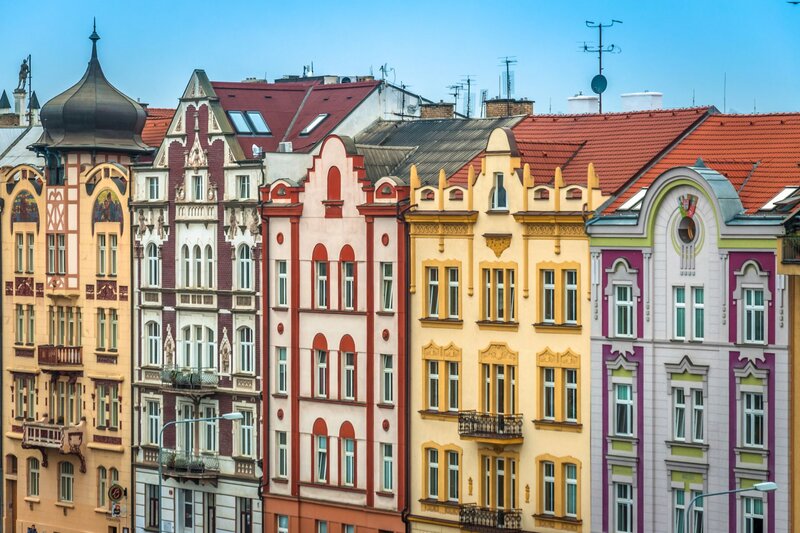 A colourful row of historic apartment buildings in Plzen features ornate facades in shades of peach, maroon, cream, yellow, and lavender, each with unique architectural details like turrets, gables, and balconies. The red-tiled rooftops and decorative moldings reflect the charm of Central European urban design under a clear blue sky.