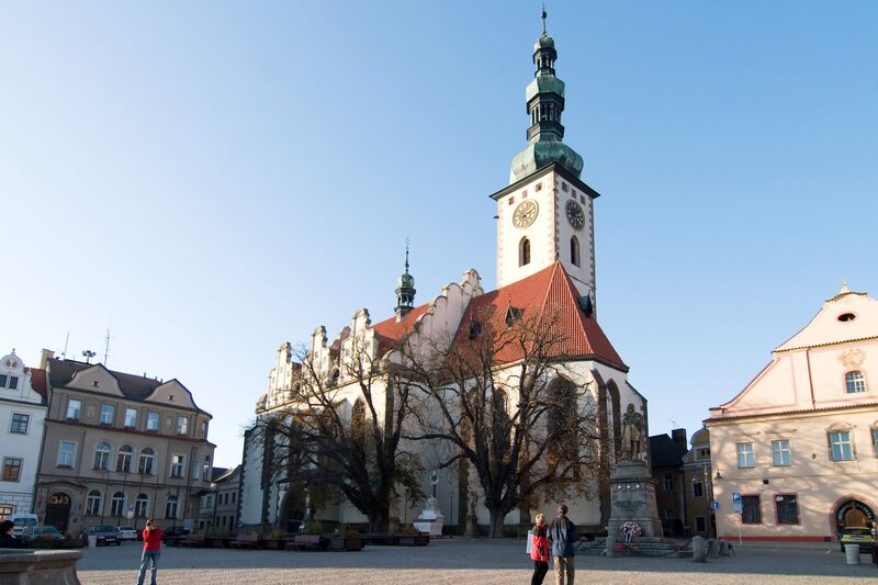 A tall church with a green copper spire and large clock tower rises above a quiet town square, surrounded by leafless trees and historic buildings. The church's red-tiled roof and white facade are illuminated by soft sunlight, while a few people walk across the open cobblestone plaza.