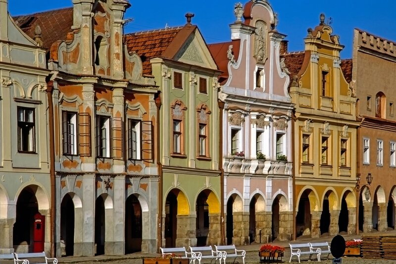 A row of pastel-coloured Renaissance and Baroque townhouses features ornate gables, arched arcades, and decorative facades in the town square of Telc, Czechia. White benches and flower boxes line the cobblestone walkway in front, creating a charming and historic atmosphere.