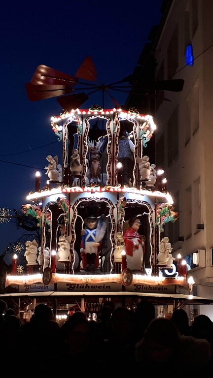 A tall, multi-tiered Christmas pyramid stall lit up at night, featuring rotating festive figures like toy soldiers, reindeer, and Santa Claus, with a glowing sign for Glühwein at the base.