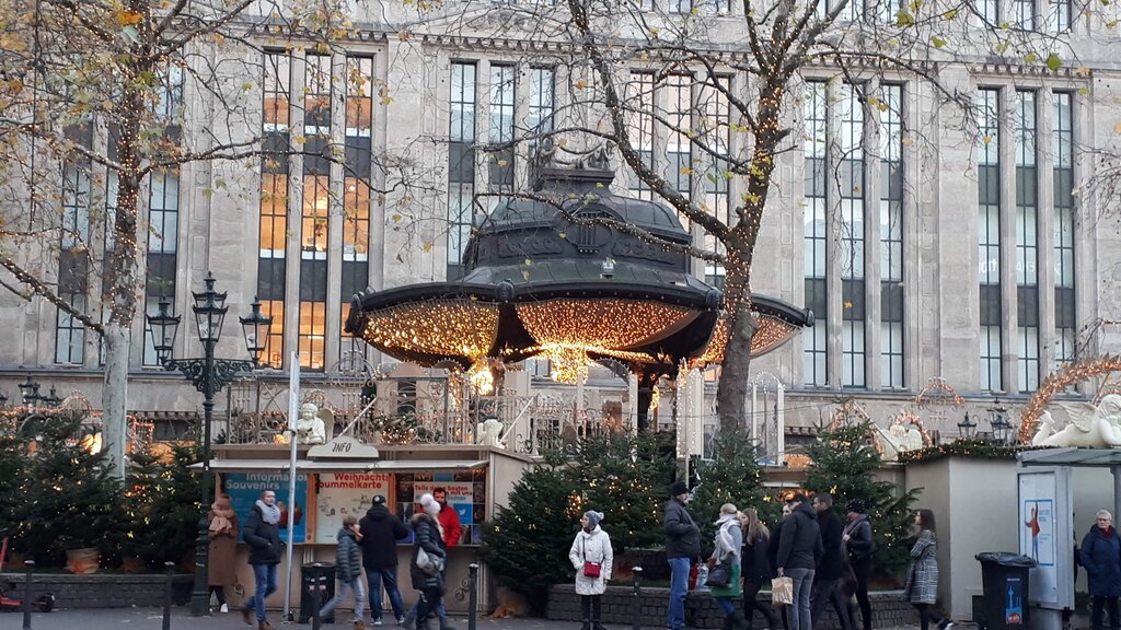 People walk past a festive wooden booth at Engelchen Markt, set beneath an ornate gazebo draped in golden Christmas lights. Surrounded by fir trees and angel decorations, the scene sits in front of a grand historic building, creating a cozy and elegant holiday atmosphere.