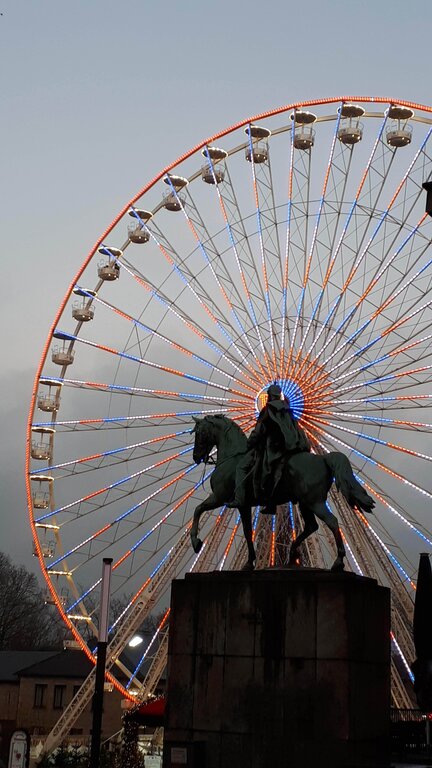 A towering Ferris wheel lit with red, white, and blue lights stands behind a bronze equestrian statue, creating a dramatic contrast against the overcast sky at the Dusseldorf Christmas Market in Germany